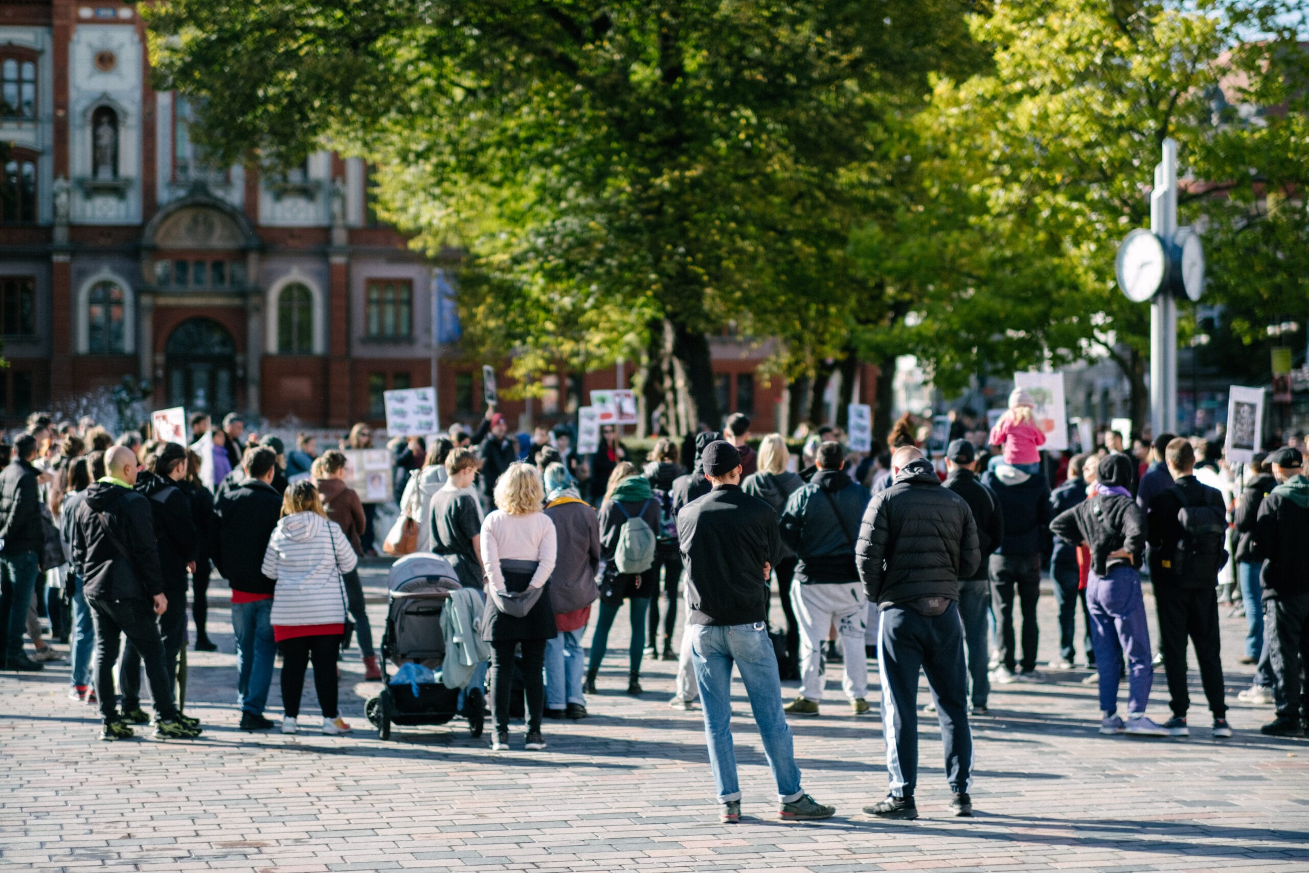 Foto von der Demo. Menschen stehen am Universitätsplatz in Rostock, einige mit Schildern. Im Hintergrund sind der Brunnen der Lebensfreude und das Hauptgebäude der Uni zu sehen. 