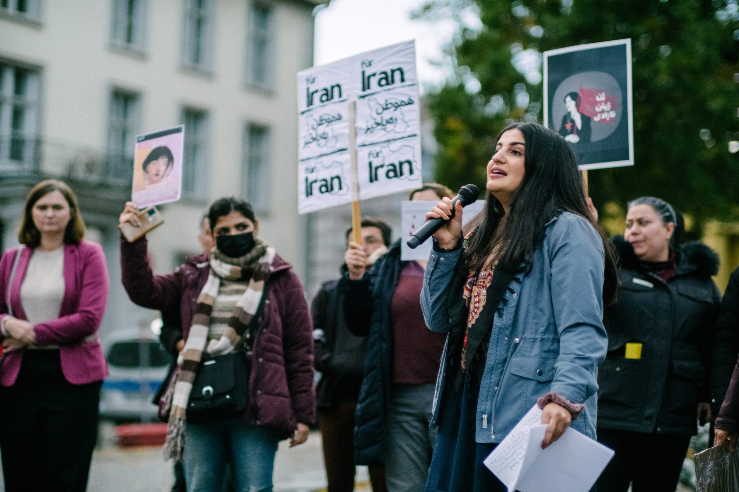 Moderatorin Esma von Women in Exile spricht in ein Mikrofon, im Hintergrund einige Menschen, die Schilder in die Höhe halten.