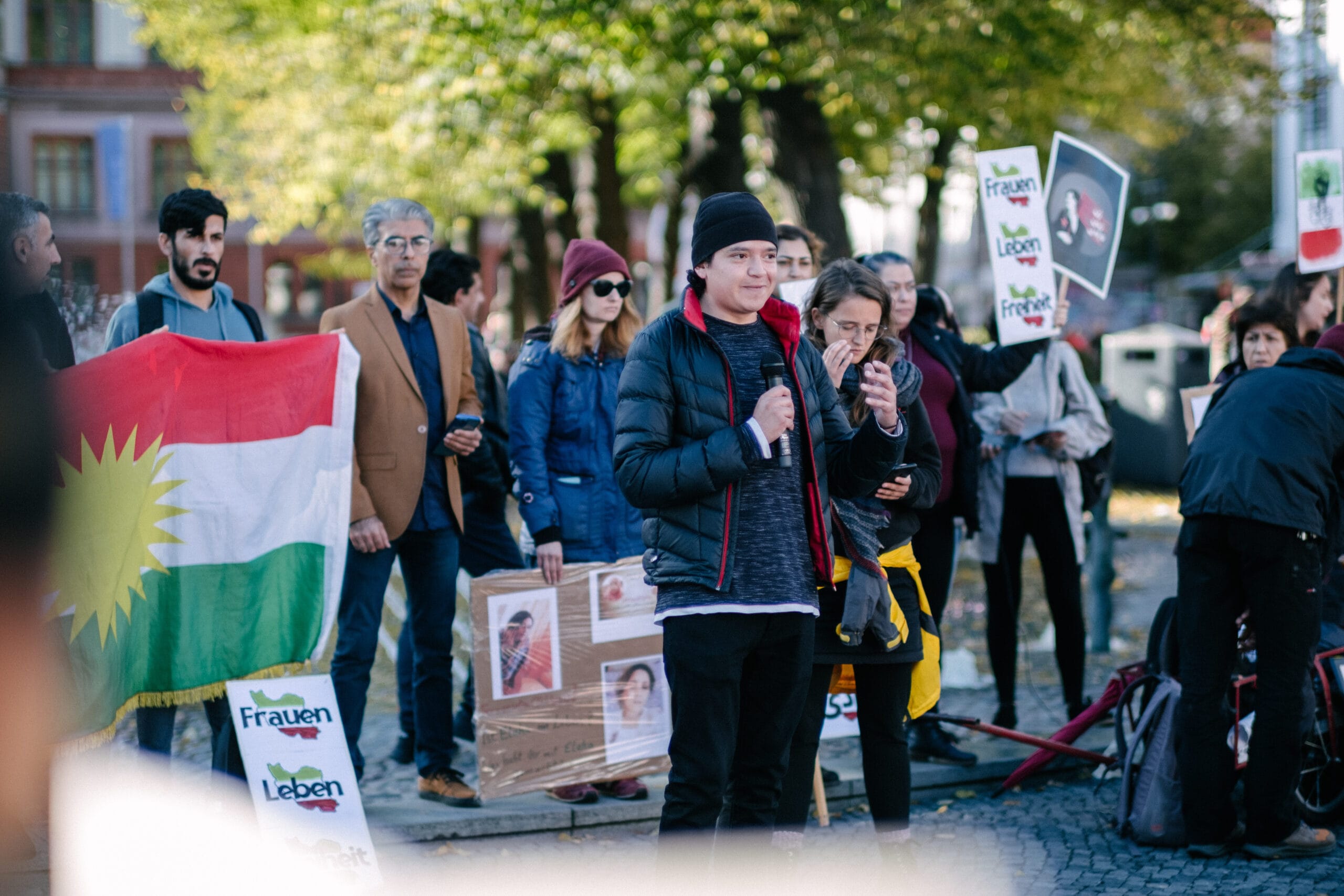 Der Freund des schwulen und christlichen Iraners, der abgeschoben werden soll, spricht in ein Mikrofon und bittet um Unterstützung. Im Hintergrund Menschen mit Schildern und einer großen Kurdistan-Flagge.