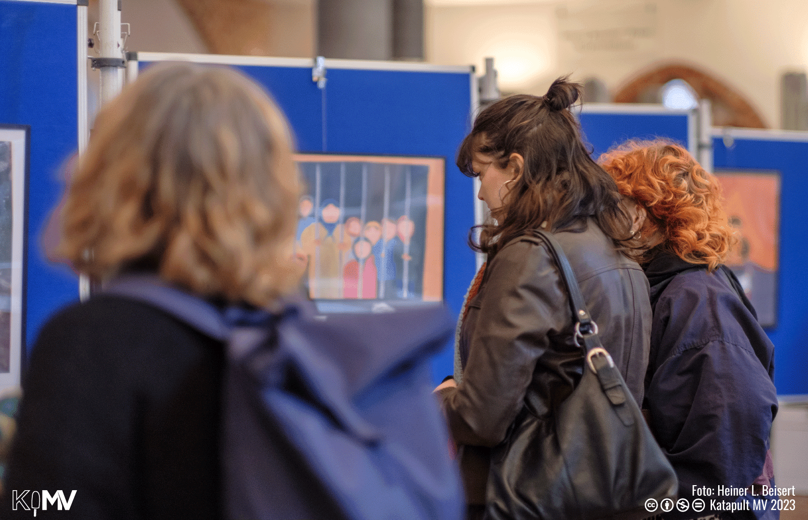 Foto von Besucher:innen, die sich die Gemälde ausschauen. Im Hintergrund ist ein Bild von Frauen und Mädchen hinter Gittern zu sehen.