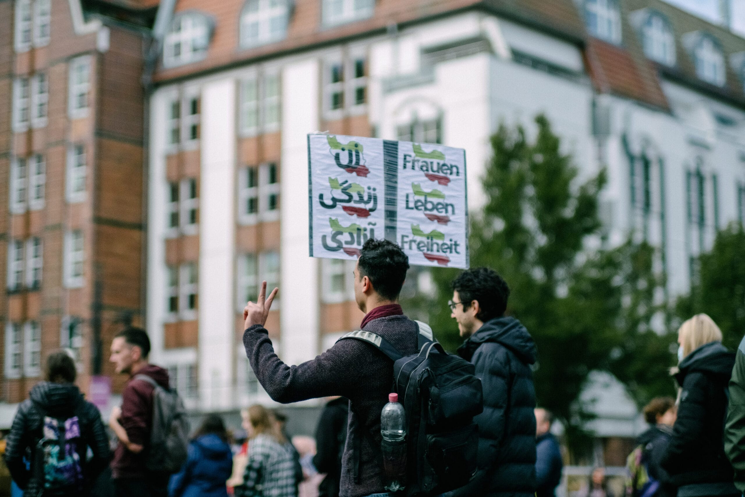 Ein Mann hält ein Schild in die Höhe mit der Aufschrift “Frauen Leben Freiheit” und formt mit Zeige- und Mittelfinger der linken Hand ein Peace-Zeichen. 