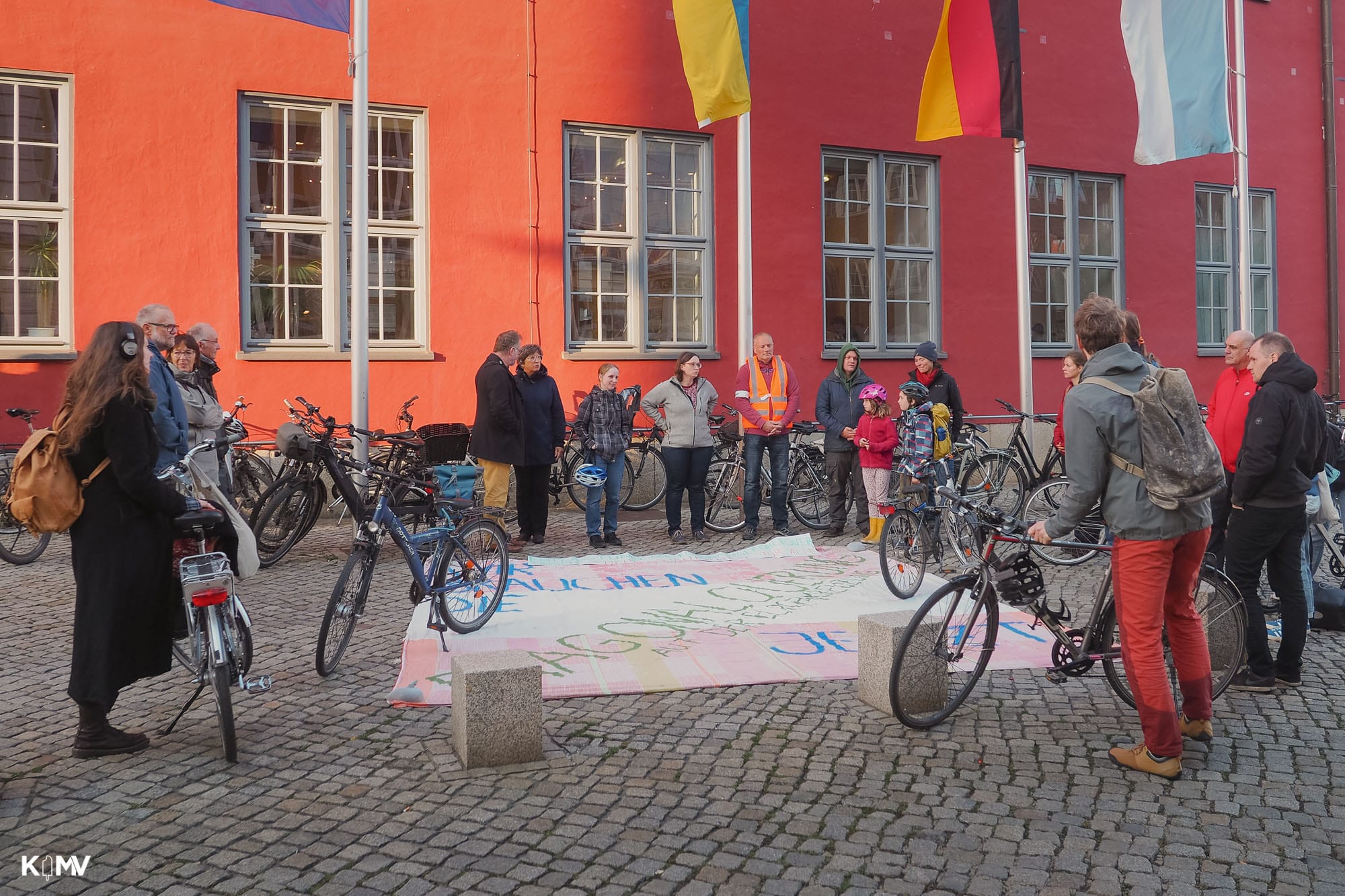 Vor dem Greifswalder Rathaus steht eine Gruppe von Menschen, einige mit ihren Fahrrädern. Sie sind um ein großes Banner versammelt, das auf dem Boden liegt. Darauf ist zu lesen: „Wir brauchen die Diagonalquerung jetzt“