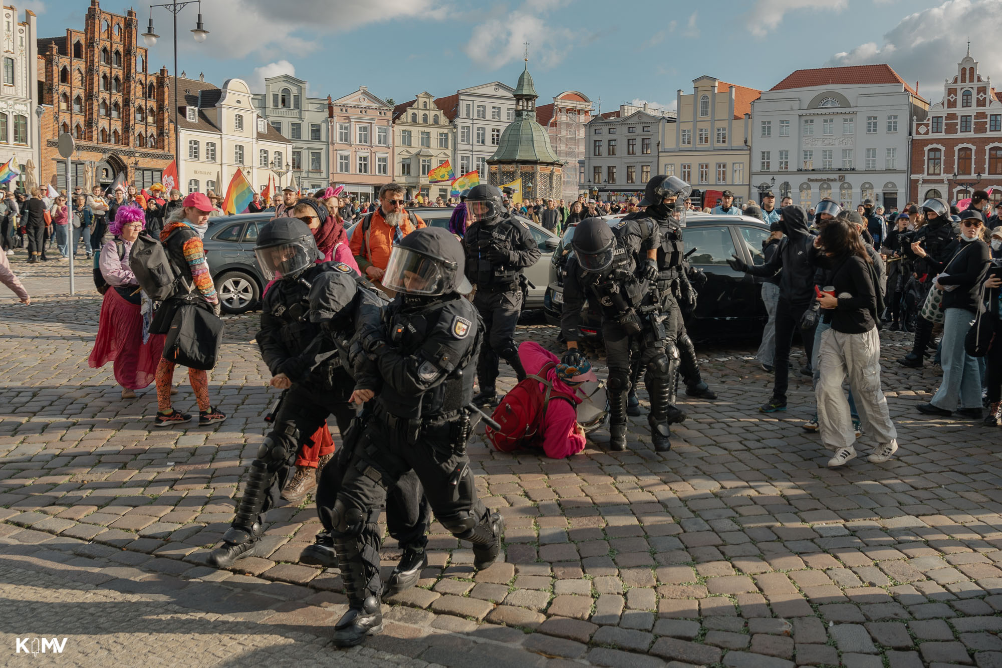 Dieses Bild zeigt eine weitere Festnahme im Rahmen des CSD in Wismar auf dem Marktplatz. Es ist zu sehen wie zwei Polizeibeamten eine schwarzgekleidete Person abführen. Im Hintergrund drückt ein weiterer Polziebeamter eine pink gekleidete Person mit Trommel gewaltvoll auf den Boden. Diese Szene ist eine Dokumentation der Polizeigewalt die teilweise gegen die Demonstrierenden stattfand. 