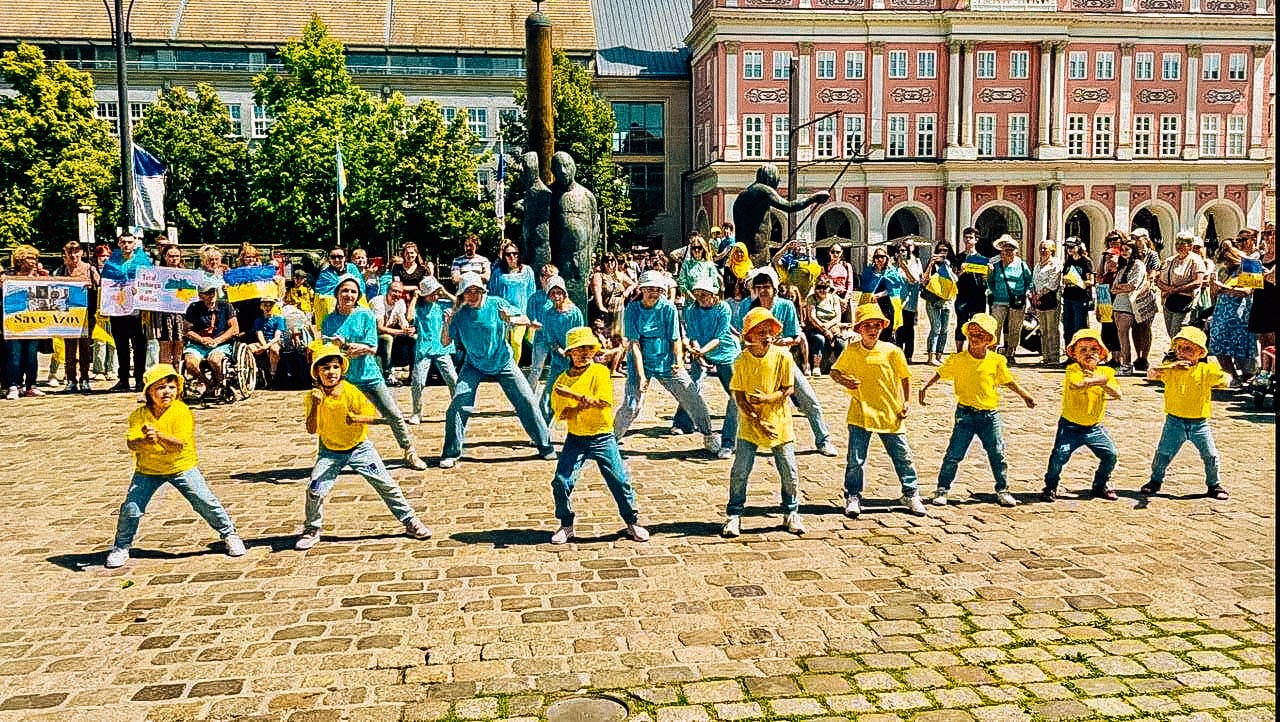 Foto; Im Vordergrund tanzen gelb und blau gekleidete Kinder. Im Hintergrund stehen Menschen mit Ukraine-Flaggen und Protestschildern.