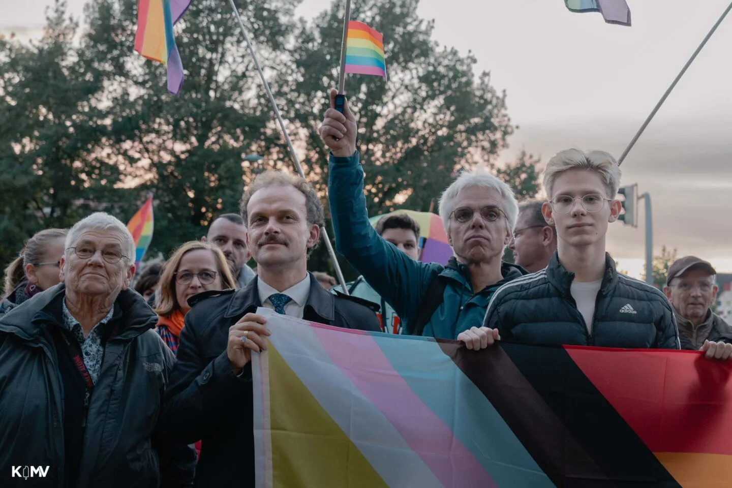 In einer Menschenmenge stehen Oberbürgermeister Silvio Witt, SPD-Stadtvertreter Michael Stieber und Mitorganisator der Demonstration Nils Berghof hinter einer Regenbogenflagge. Im Hintergrund halten Menschen kleinere Flaggen hoch.