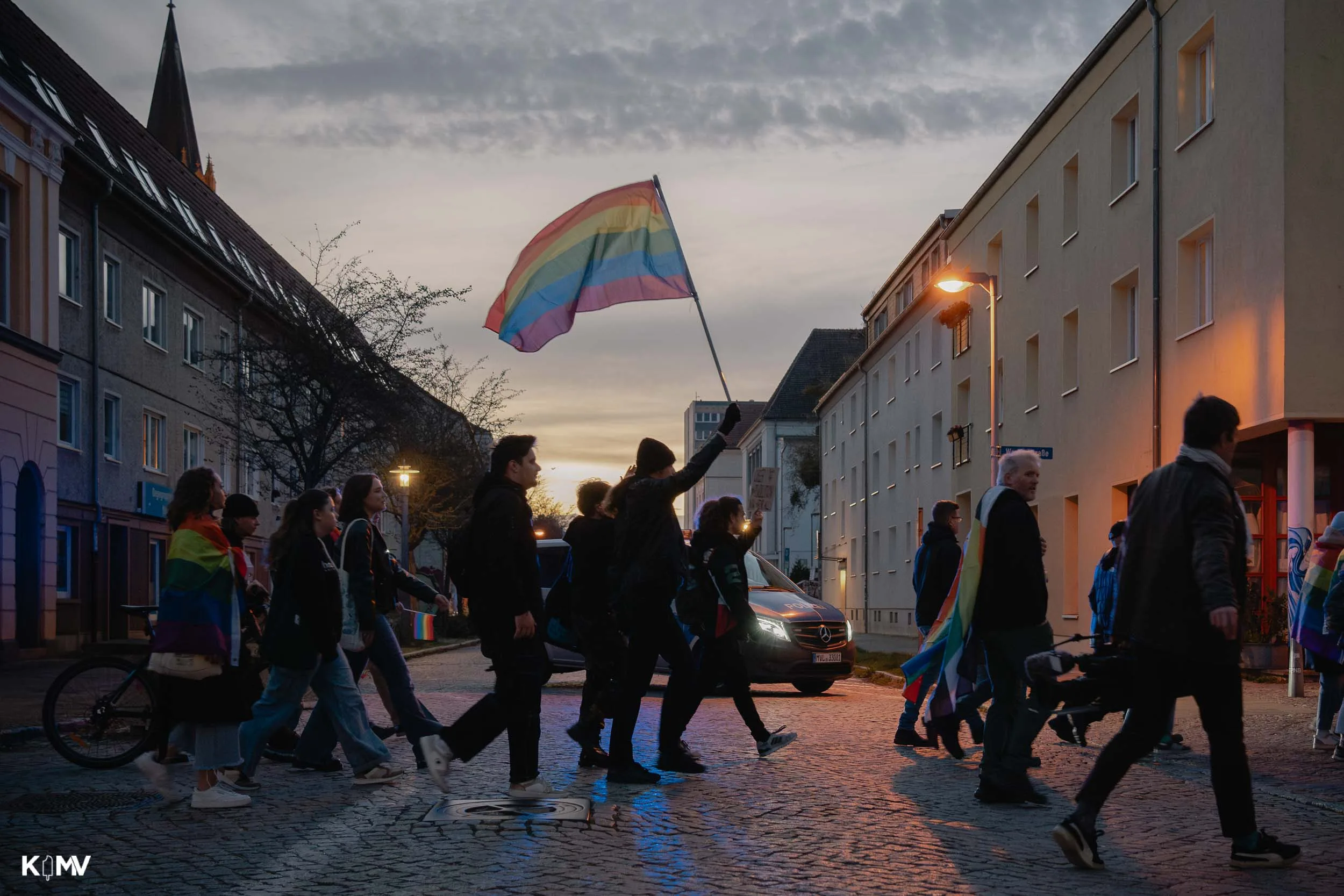 In der Dämmerung kommen Menschen der Demo an einer Straßenecke vorbei und halten eine große Regenbogenflagge hoch. 