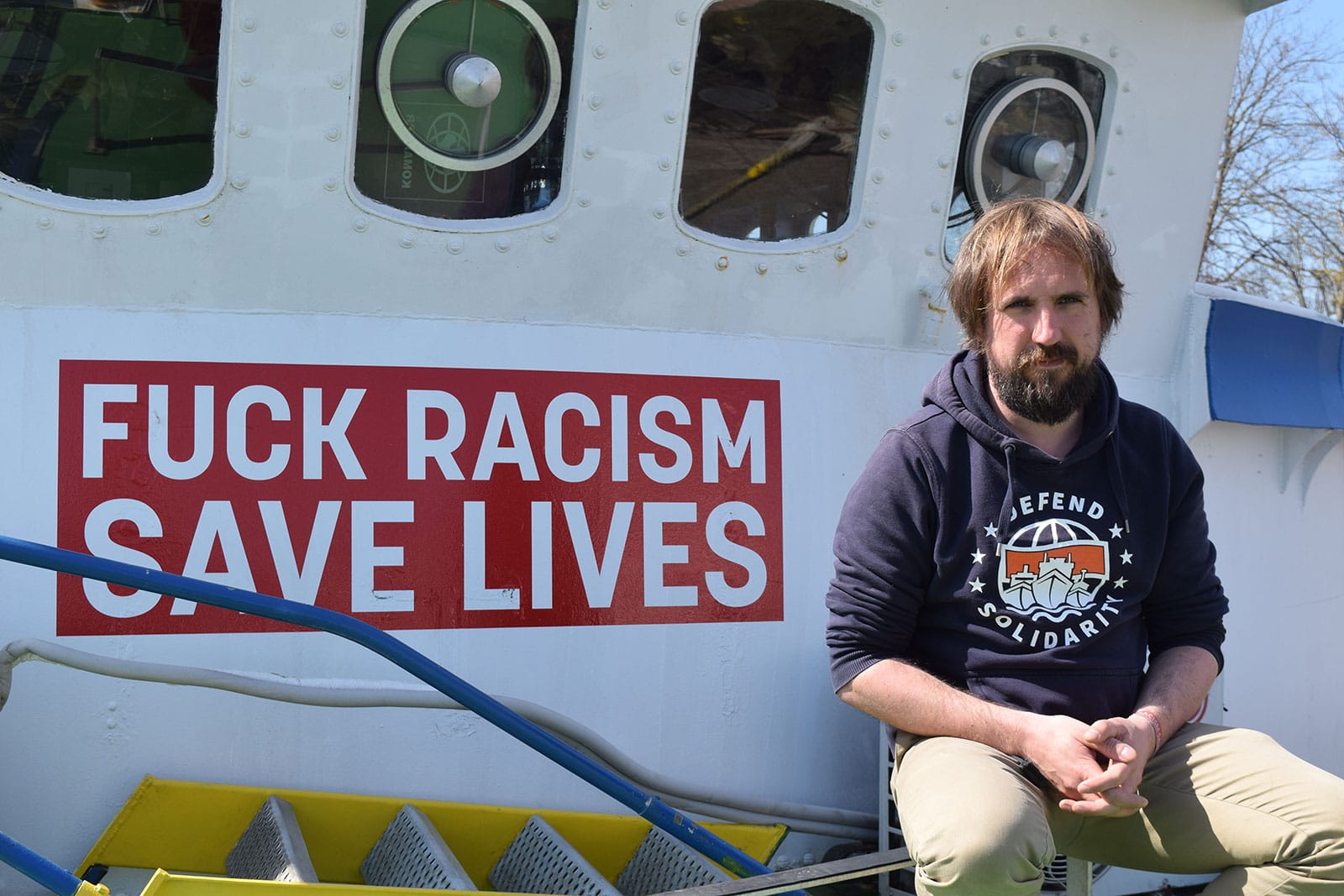 Eine Person sitzt neben dem Schriftzug „Fuck Racism – Save Lives“, der auf die Brücke eines Schiffs gemalt wurde.