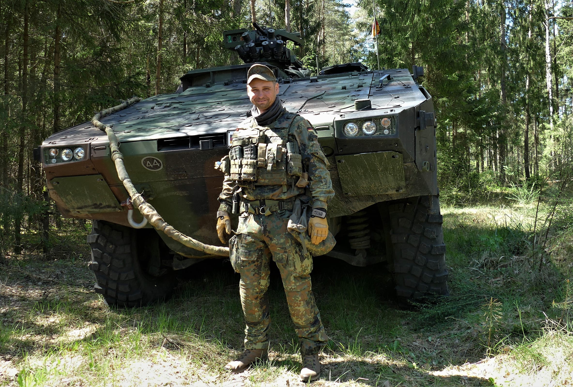 Mann in Flecktarnanzug steht vor einem Panzerfahrzeug.