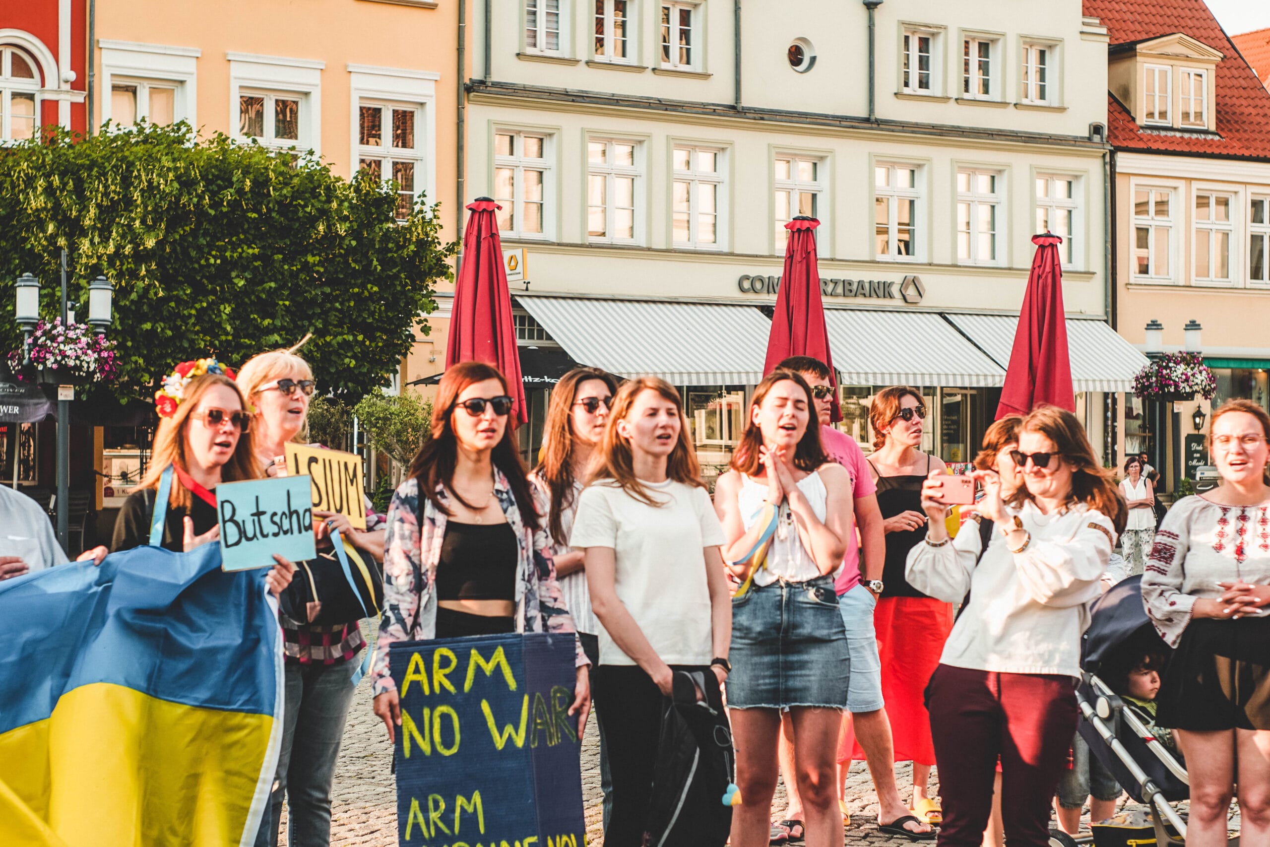 Foto; Demonstrant:innen in sommerlicher Kleidung sind beim Singen abgebildet. Sie tragen u.a. eine Ukraine-Flagge und ein 